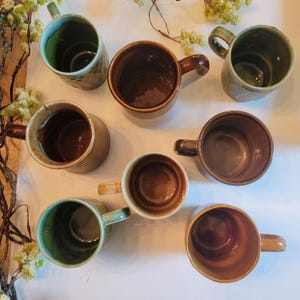May include: A collection of handmade ceramic mugs in various earth-tone colors, including brown, green, and beige. The mugs have unique textures and designs, with handles for easy gripping. Some mugs have a ribbed design, while others feature embossed patterns.
