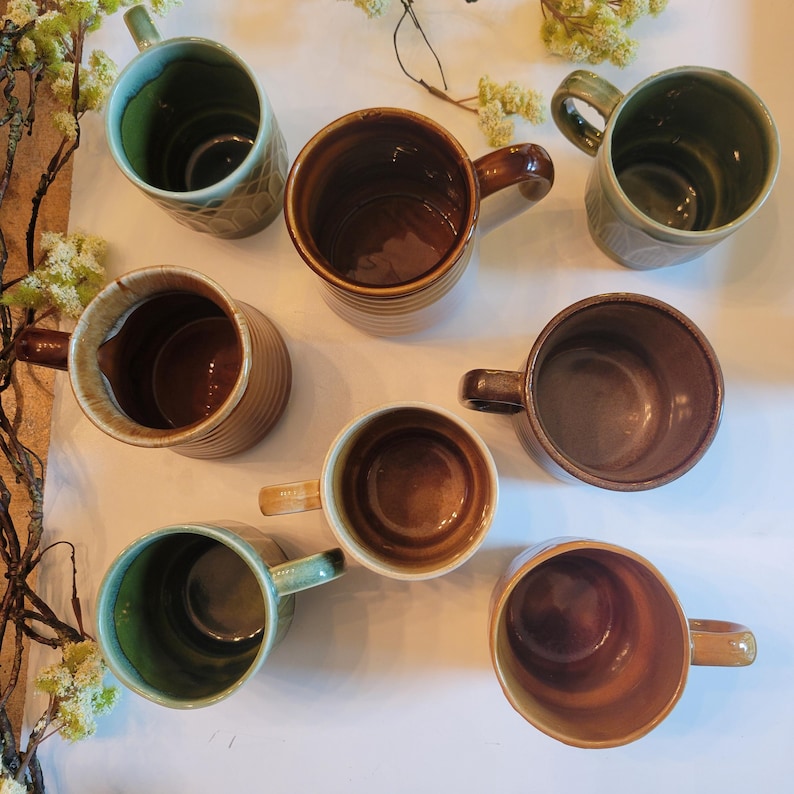 May include: A collection of handmade ceramic mugs in various earth-tone colors, including brown, green, and beige. The mugs have unique textures and designs, with handles for easy gripping. Some mugs have a ribbed design, while others feature embossed patterns.