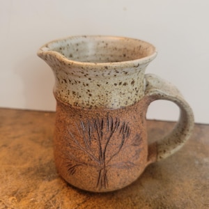 May include: A handcrafted ceramic jug with a light beige speckled rim and a brown body. The body features an embossed tree design. The jug has a handle and a spout for easy pouring. The pottery has a rustic, earthy aesthetic.