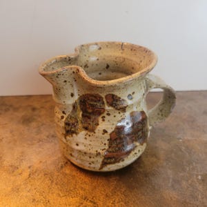 May include: A handmade ceramic jug with a distinctive, natural form. The jug has a speckled beige glaze with brown and dark brown abstract patterns. It features a curved handle and a spout. A rustic, functional piece, ideal for serving.