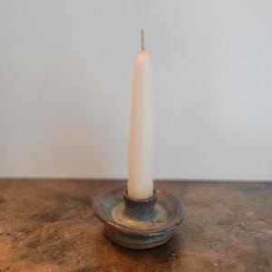 Peut inclure: Un bougeoir en céramique avec une bougie crème. Le bougeoir est une petite coupelle ronde avec un centre surélevé, verni dans des tons de bleu et de vert. La bougie est haute et élancée, avec une mèche visible.