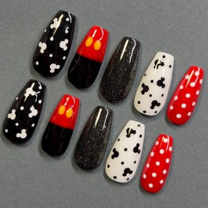 May include: A set of ten artificial nails in various designs. The nails feature black and white Mickey Mouse patterns, red and black Mickey Mouse designs, black glitter, and red nails with white polka dots. The nails are oval-shaped.