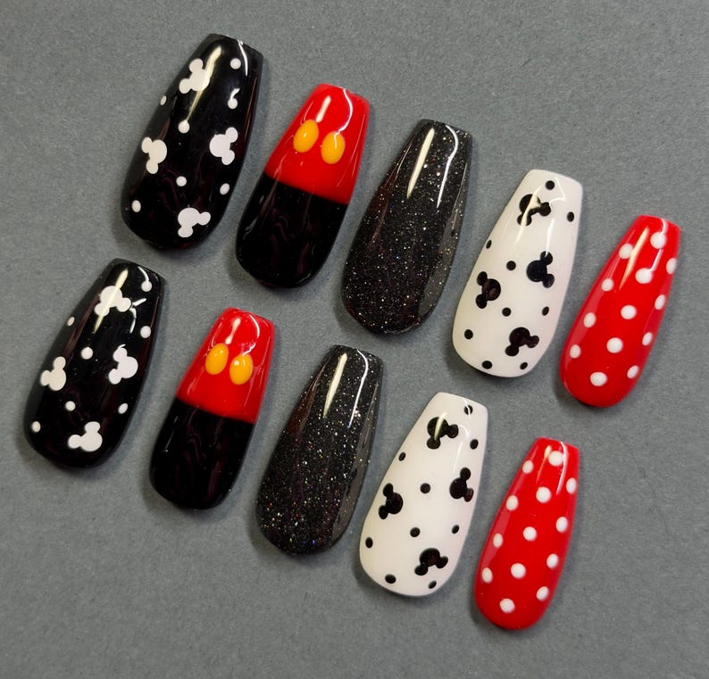 May include: A set of ten artificial nails in various designs. The nails feature black and white Mickey Mouse patterns, red and black Mickey Mouse designs, black glitter, and red nails with white polka dots. The nails are oval-shaped.