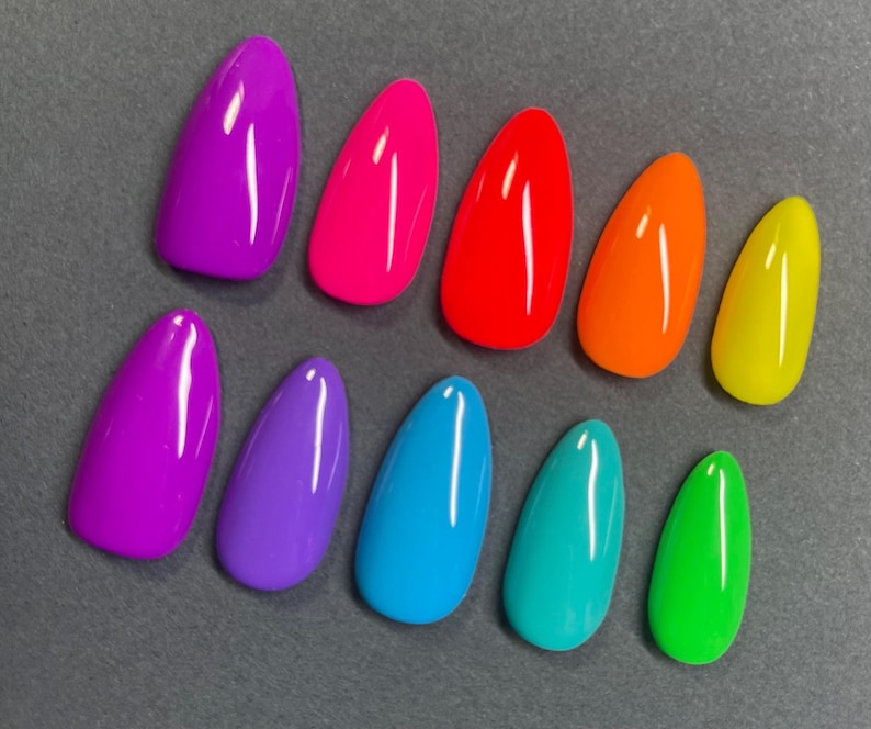 May include: A collection of ten brightly colored, almond-shaped artificial nails. The colors include shades of purple, pink, red, orange, yellow, blue, teal, and green. The nails are arranged on a gray surface, showcasing a vibrant spectrum of hues.