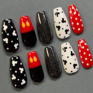 May include: A collection of ten artificial nails in various designs. The nails feature black and white Mickey Mouse designs, red and black Mickey Mouse designs, black glitter, and red nails with white polka dots. The nails are arranged on a gray surface.