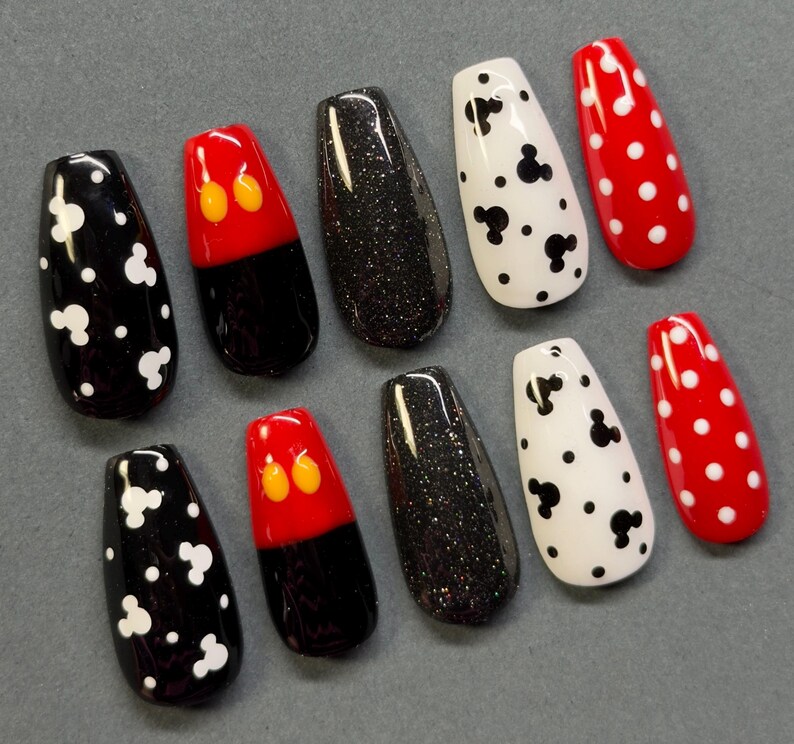 May include: A collection of ten artificial nails in various designs. The nails feature black and white Mickey Mouse designs, red and black Mickey Mouse designs, black glitter, and red nails with white polka dots. The nails are arranged on a gray surface.