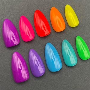 May include: A collection of ten glossy, almond-shaped artificial nails in a variety of vibrant colors. The colors include purple, hot pink, red, orange, yellow, blue, teal, and green. The nails are arranged on a dark gray surface.