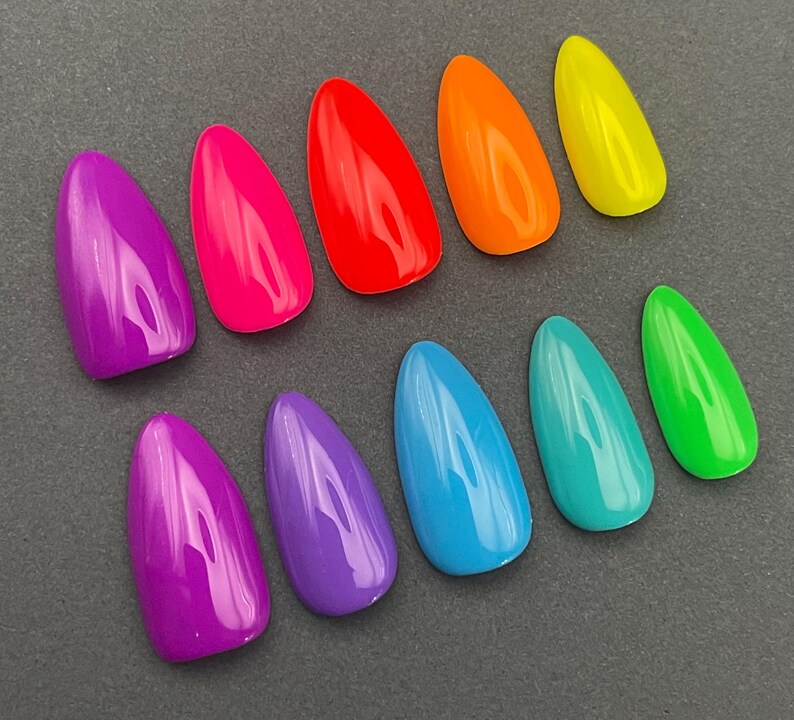May include: A collection of ten glossy, almond-shaped artificial nails in a variety of vibrant colors. The colors include purple, hot pink, red, orange, yellow, blue, teal, and green. The nails are arranged on a dark gray surface.