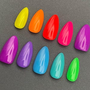 May include: A collection of ten brightly colored, glossy, artificial nails. The nails are in a variety of vibrant colors, including yellow, orange, red, pink, purple, blue, teal, and green. The nails are arranged on a dark gray surface.