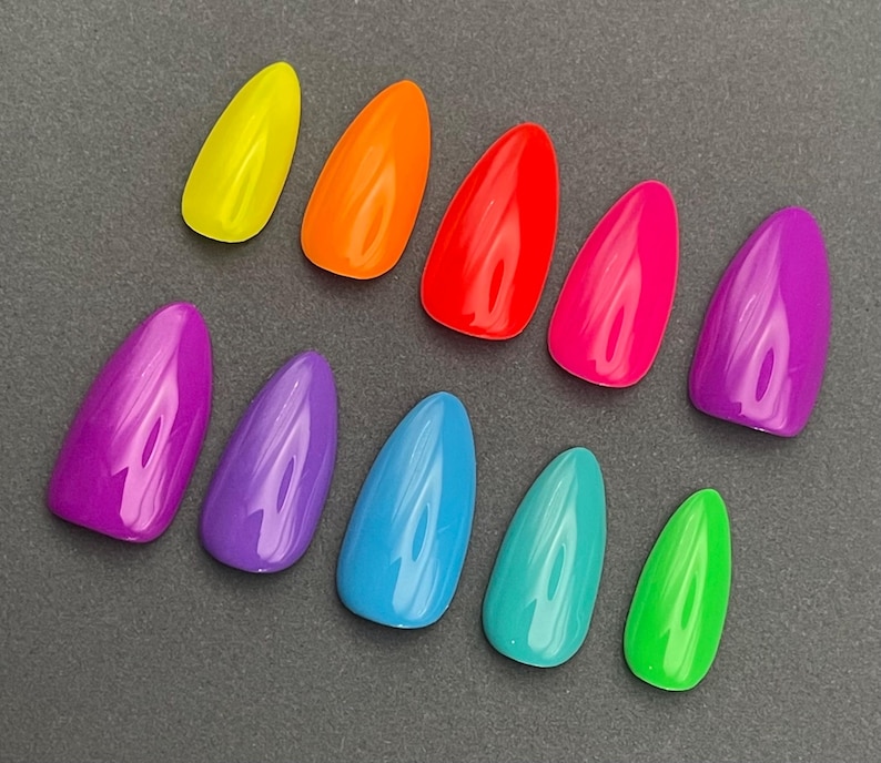 May include: A collection of ten brightly colored, glossy, artificial nails. The nails are in a variety of vibrant colors, including yellow, orange, red, pink, purple, blue, teal, and green. The nails are arranged on a dark gray surface.
