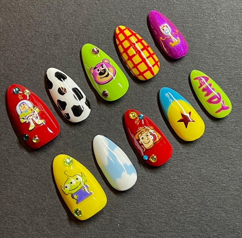 Op de afbeelding: Een set van tien amandelvormige kunstnagels met verschillende ontwerpen. De ontwerpen omvatten stripfiguren, een koeienprint en het woord "Andy". Kleuren zijn rood, geel, groen, wit, blauw en paars. Sommige nagels hebben strass-steentjes.