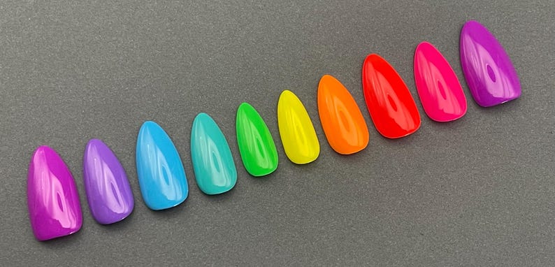 May include: A collection of ten glossy, almond-shaped artificial nails in a rainbow of colors. The colors include purple, blue, teal, green, yellow, orange, red, pink, and another shade of purple. The nails are arranged in a diagonal line.