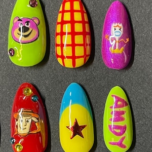 Op de afbeelding: Set van zes amandelvormige persnagels met cartoonkarakterontwerpen. De nagels bevatten personages als Lotso, Woody en Forky, met een geel en rood rasterpatroon en de naam "Andy".
