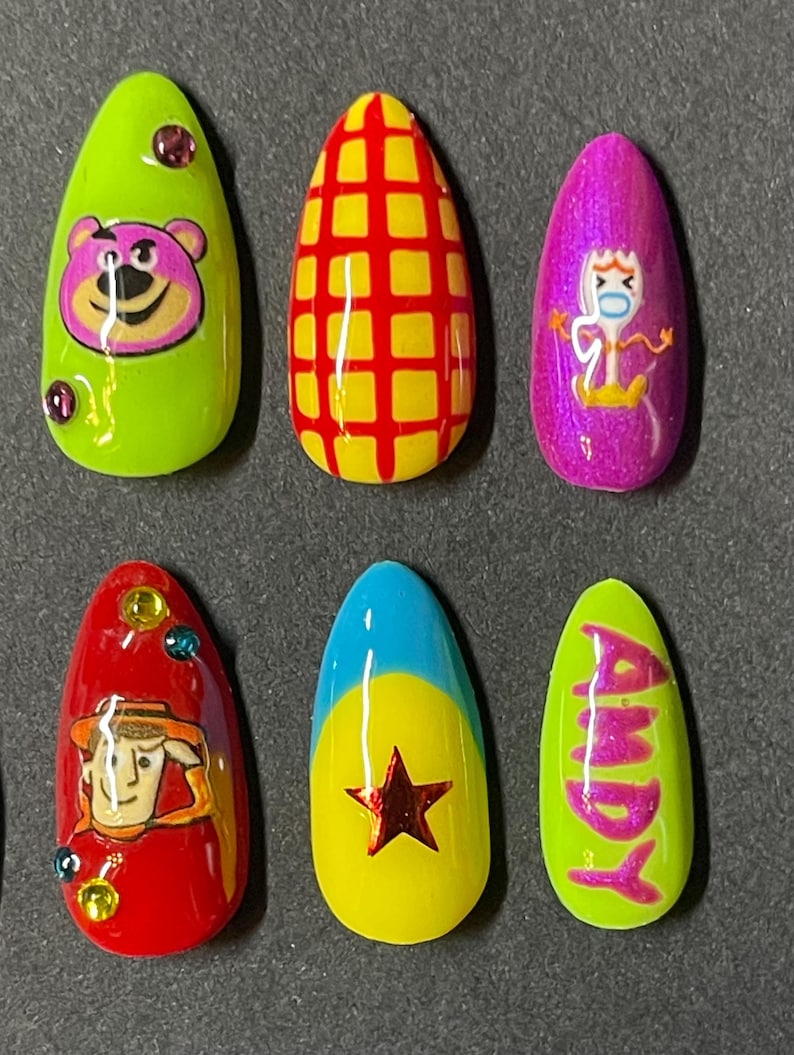 Op de afbeelding: Set van zes amandelvormige persnagels met cartoonkarakterontwerpen. De nagels bevatten personages als Lotso, Woody en Forky, met een geel en rood rasterpatroon en de naam "Andy".