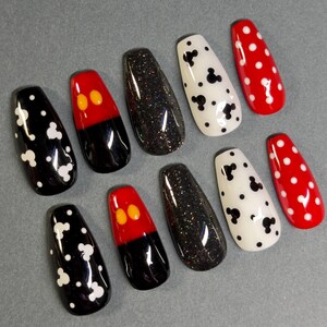 May include: A set of ten artificial nails with various designs. The nails feature black, red, and white colors. Some nails have white Mickey Mouse head shapes, while others have red and black designs with yellow accents. Other nails are black with glitter or white with black Mickey Mouse shapes.