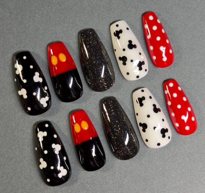 May include: A set of ten artificial nails with various designs. The nails feature black, red, and white colors. Some nails have white Mickey Mouse head shapes, while others have red and black designs with yellow accents. Other nails are black with glitter or white with black Mickey Mouse shapes.