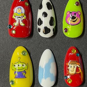 Op de afbeelding: Set van zes opzetnagels met cartoonkarakterontwerpen. De nagels bevatten personages als Buzz Lightyear, een koeienprint, Lotso, een alien, een wolkenontwerp en Woody. Elke nagel heeft een andere kleur en heeft decoratieve edelstenen.