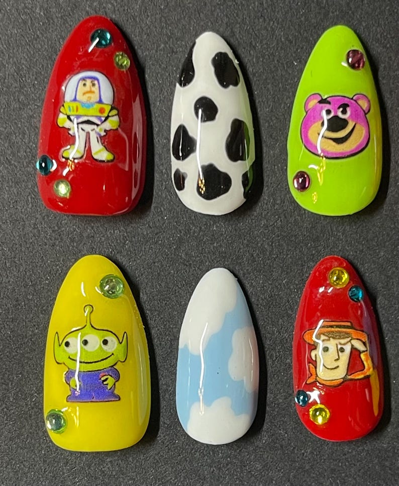 Op de afbeelding: Set van zes opzetnagels met cartoonkarakterontwerpen. De nagels bevatten personages als Buzz Lightyear, een koeienprint, Lotso, een alien, een wolkenontwerp en Woody. Elke nagel heeft een andere kleur en heeft decoratieve edelstenen.