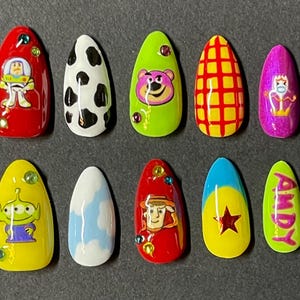 Op de afbeelding: Een set van tien kunstnagels met verschillende ontwerpen ge&iuml;nspireerd op de animatiefilm Toy Story. De nagels tonen personages als Buzz Lightyear, Lotso en Woody, samen met andere thema-patronen. Elke nagel heeft een andere kleur, waaronder rood, geel en groen.