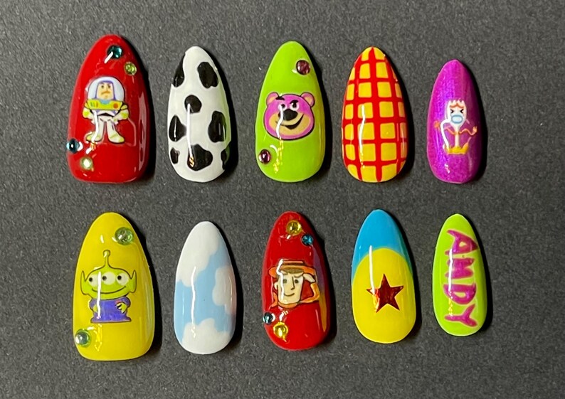 Op de afbeelding: Een set van tien kunstnagels met verschillende ontwerpen ge&iuml;nspireerd op de animatiefilm Toy Story. De nagels tonen personages als Buzz Lightyear, Lotso en Woody, samen met andere thema-patronen. Elke nagel heeft een andere kleur, waaronder rood, geel en groen.