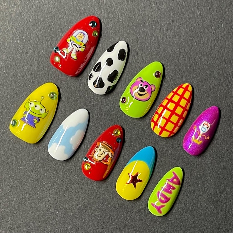 Op de afbeelding: Een set van tien amandelvormige kunstnagels met verschillende ontwerpen ge&iuml;nspireerd op de animatiefilm Toy Story. De nagels tonen personages als Buzz Lightyear en Woody, samen met andere thema-patronen en de naam "ANDY".