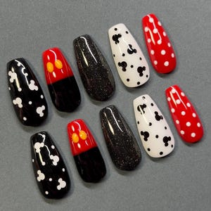 May include: A set of ten artificial nails in various designs. The nails feature black and white polka dots, black glitter, red with white polka dots, and red with yellow and orange accents. The nails are coffin-shaped.