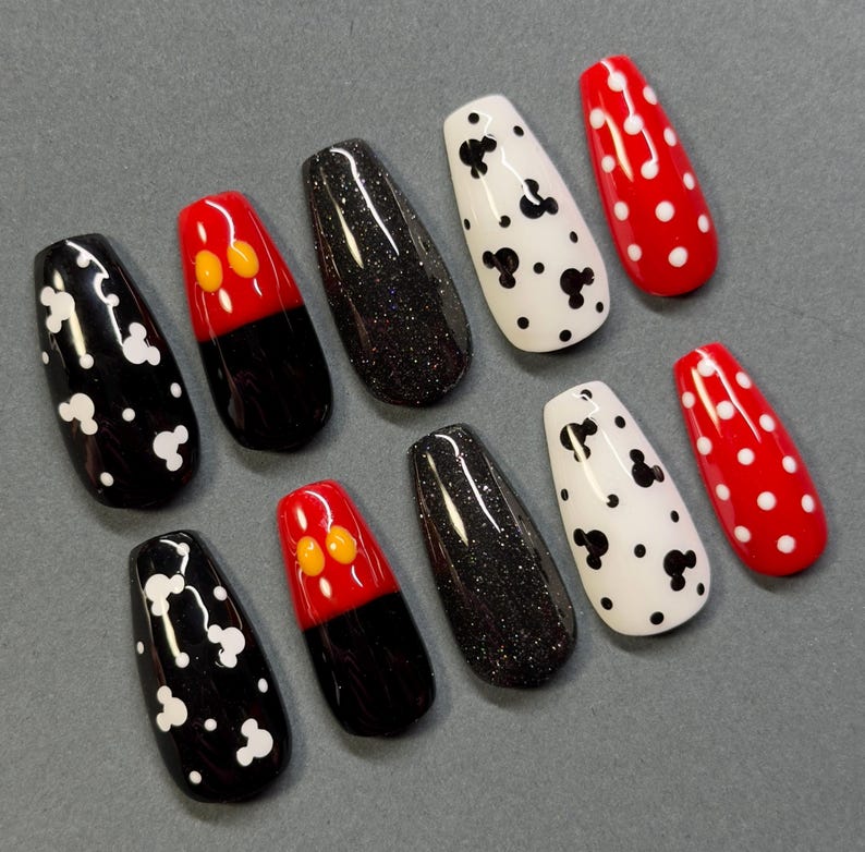 May include: A set of ten artificial nails in various designs. The nails feature black and white polka dots, black glitter, red with white polka dots, and red with yellow and orange accents. The nails are coffin-shaped.