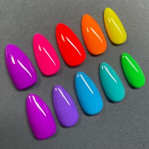 May include: A collection of ten brightly colored, oval-shaped artificial nails. The nails are arranged on a gray surface and feature a glossy finish. Colors include purple, pink, red, orange, yellow, teal, blue, and green.