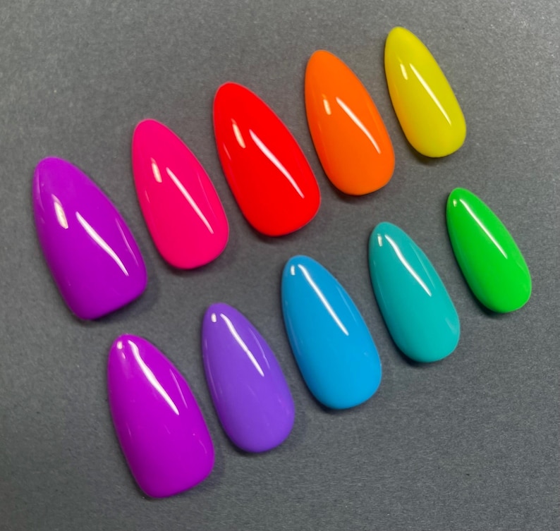 May include: A collection of ten brightly colored, oval-shaped artificial nails. The nails are arranged on a gray surface and feature a glossy finish. Colors include purple, pink, red, orange, yellow, teal, blue, and green.