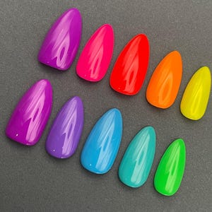May include: A collection of ten brightly colored, glossy, almond-shaped artificial nails. The colors include shades of purple, pink, red, orange, yellow, blue, teal, and green. The nails are arranged on a dark gray surface.