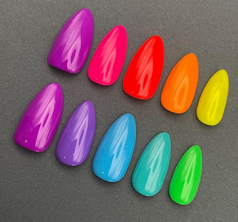 May include: A collection of ten brightly colored, glossy, almond-shaped artificial nails. The colors include shades of purple, pink, red, orange, yellow, blue, teal, and green. The nails are arranged on a dark gray surface.