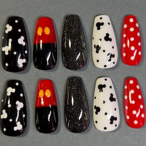 May include: A set of ten artificial nails in various designs. The designs include black nails with white Mickey Mouse shapes, red and black nails with yellow Mickey Mouse ears, black glitter nails, white nails with black Mickey Mouse shapes, and red nails with white polka dots.