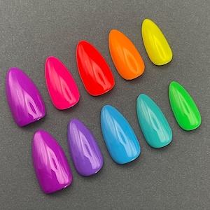 May include: A collection of ten brightly colored, almond-shaped artificial nails. The nails are arranged on a gray surface and feature a glossy finish in shades of purple, pink, red, orange, yellow, green, teal, and blue.