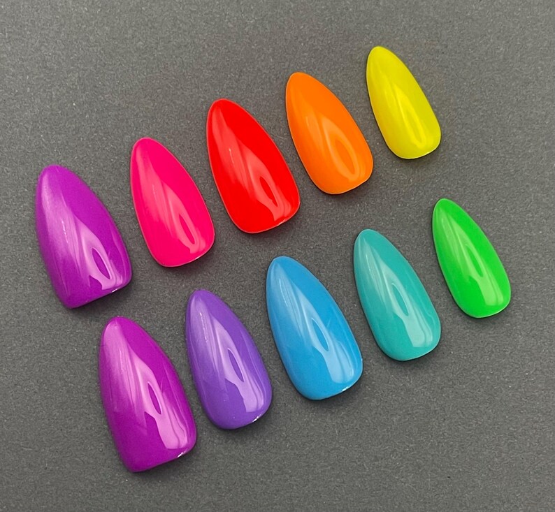 May include: A collection of ten brightly colored, almond-shaped artificial nails. The nails are arranged on a gray surface and feature a glossy finish in shades of purple, pink, red, orange, yellow, green, teal, and blue.