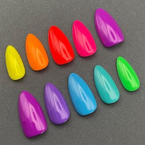 May include: A collection of ten brightly colored, almond-shaped artificial nails. The colors include yellow, orange, red, hot pink, purple, blue, teal, and green. The nails are arranged on a dark gray surface.