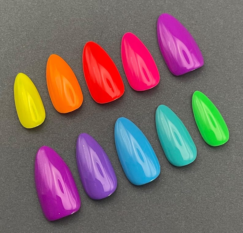 May include: A collection of ten brightly colored, almond-shaped artificial nails. The colors include yellow, orange, red, hot pink, purple, blue, teal, and green. The nails are arranged on a dark gray surface.