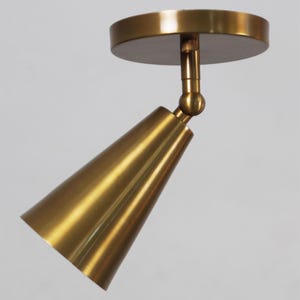 May include: A gold-colored, cone-shaped spotlight fixture with a round base. The spotlight is attached to the base with a ball joint, allowing for adjustable positioning. The fixture is made of metal and has a polished finish.