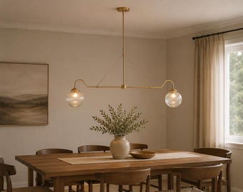 Puede incluir: Una lámpara de araña lineal con acabado en latón que presenta dos pantallas esféricas de cristal estriado suspendidas de una barra horizontal central. El accesorio se sostiene mediante una varilla vertical y cables finos.