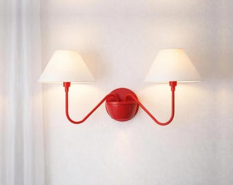 May include: A red wall sconce with two white lampshades. The sconce has a curved design with a small bowl in the center. The lampshades are conical and the fixture is mounted on a white wall.