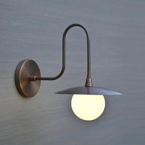 May include: A bronze-colored wall-mounted light fixture. It features a circular base, a curved arm, and a downward-facing, saucer-shaped shade. A spherical, glowing light bulb is visible below the shade. The fixture is mounted on a gray wall.