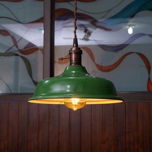 Puede incluir: Una lámpara colgante verde con un accesorio de latón y una bombilla cálida y brillante. La lámpara tiene una pantalla redondeada en forma de cuenco y un cable marrón. El fondo presenta una pared de madera y una ventana con vidrieras de colores.