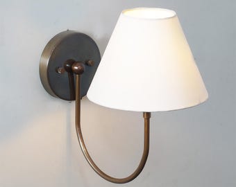May include: A wall-mounted lamp with a white lampshade and a bronze-colored metal frame. The lampshade is cone-shaped, and the frame has a curved arm. The lamp is designed to be mounted on a wall and provides ambient lighting.