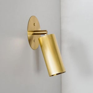 Può includere: Applique da parete moderna color oro. L'apparecchio presenta una base circolare, una luce cilindrica e un braccio regolabile. L'applique è montata su una parete grigio chiaro, offrendo un'estetica minimalista.
