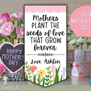 Puede incluir: Un cartel del Día de la Madre con el texto "Mothers plant the seeds of love that grow forever". Una pizarra en forma de corazón dice "Happy Mother's Day". Flores rosas y blancas en el fondo.