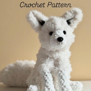 Op de afbeelding: Een witte gehaakte poolvos met zwarte ogen en neus, en grote, rechtopstaande oren. De tekst "Arctic Fox Crochet Pattern" staat boven de vos. De vos zit op een wit oppervlak tegen een lichtgele achtergrond.