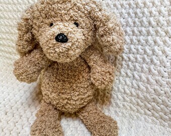 May include: A plush, brown dog toy with a curly texture. The stuffed animal has a black nose and small, dark eyes. The dog is sitting on a white, textured surface.