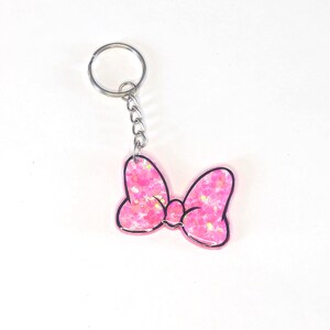 May include: A pink bow-shaped keyring with a glittery design and a black outline. The keyring has a silver metal ring and chain. The bow is a bright pink colour with glitter accents.