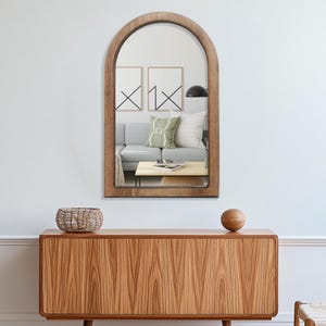 May include: A wooden arched mirror with a natural finish hangs on a white wall. The mirror reflects a living room scene with a gray sofa, coffee table, and artwork. Below, a wooden cabinet with a woven basket and a wooden sphere.