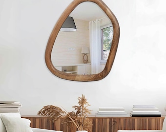 May include: A modern, irregularly shaped mirror with a natural wood frame. The mirror hangs on a white wall, reflecting a room with a window and a lamp. The frame has a smooth, organic shape.