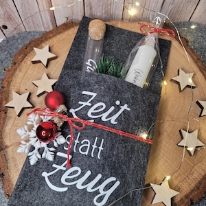 Könnte beinhalten: Eine dunkelgraue Filz-Geschenktasche mit den Worten "Zeit statt Zeug" in Weiß. Die Tasche ist mit rotem Faden gebunden und mit einem roten Ornament und einem Schneeflocke verziert. Ein Glasröhrchen mit Korken und eine weiße Kerze sind darin.